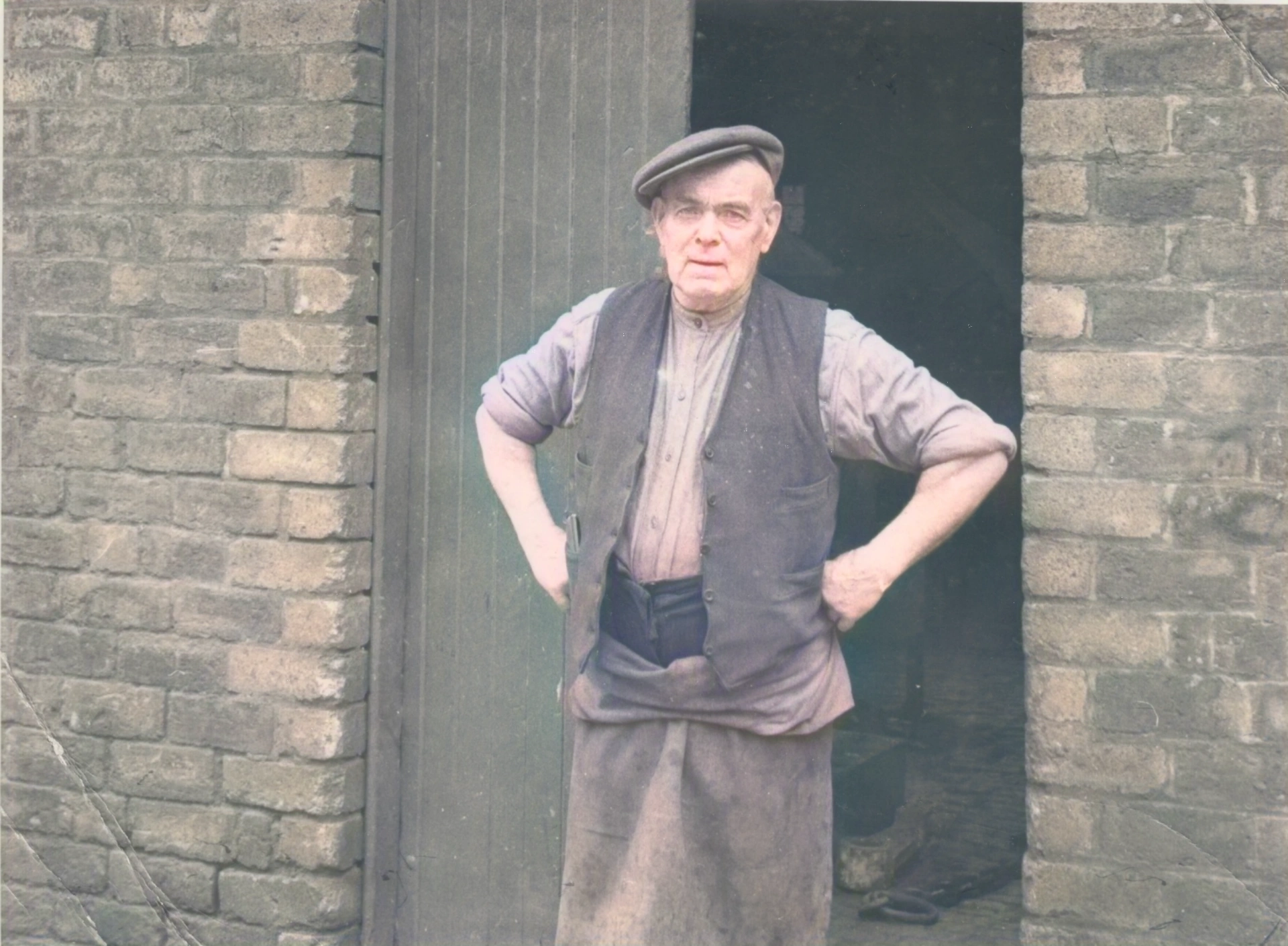 A blacksmith in work clothes standing in the doorway of the forge building